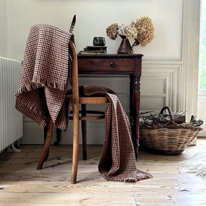 May include: A brown and black houndstooth patterned throw blanket draped over a wooden chair. The blanket has a fringed edge. A small wooden side table with a vase of dried flowers and a basket of logs are in the background.