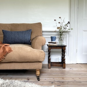 May include: A tan linen sofa with a dark blue throw pillow and a burnt orange throw blanket. A small wooden side table with a blue mug and a vase of dried flowers sits next to the sofa.