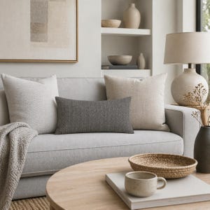 Op de afbeelding: Een woonkamer met een lichtgrijze bank versierd met kussens in neutrale tinten. Een grijs visgraat lendenkussen is centraal geplaatst. Een ronde houten salontafel toont een geweven dienblad, een keramische mok en een stapel boeken.