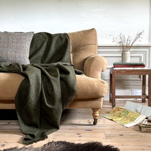 Puede incluir: Un sofá beige con una manta verde y un cojín a cuadros. Una pequeña mesa auxiliar de madera con libros y un jarrón con tallos secos. Un mapa está en el suelo. Ambiente acogedor.