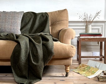 May include: A cozy living room scene featuring a tan sofa with a green blanket draped over it. A plaid pillow rests on the sofa. A small wooden side table holds books and a vase with dried stems. A map is on the floor.