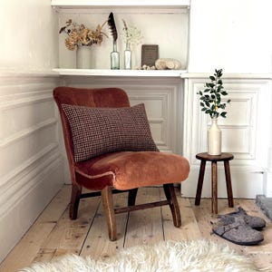 May include: A burnt orange velvet armchair with a checkered patterned cushion. The chair has wooden legs and is sitting on a wooden floor. A small wooden stool with a white vase and green plant is in the background.