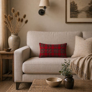 Op de afbeelding: Een beige bank met een rood geruit lendenkussen. Een beige sierkussen en een deken met franjes liggen op de bank. Een houten bijzettafel met een vaas met gedroogde planten en een kleine kom staat links. Een houten salontafel met een kom en een kleine plant staat ervoor.