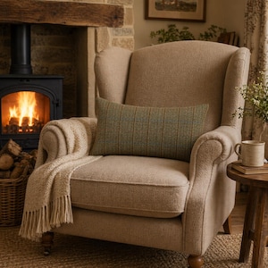 Op de afbeelding: Een beige fauteuil met een groen en bruin gestreept lendenkussen. Een crèmekleurige deken met franjes is over de arm gedrapeerd. Een kleine houten bijzettafel met een mok. Een open haard met een brandend vuur op de achtergrond.