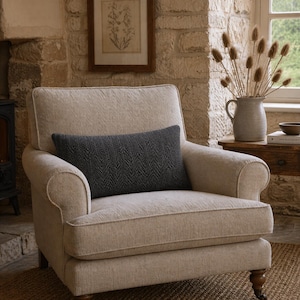 Op de afbeelding: Een beige gestoffeerde fauteuil met een donkergrijs kussen met visgraatpatroon. De stoel heeft gerolde armleuningen en houten poten. Een ingelijste botanische print hangt aan de stenen muur. Een vaas met gedroogde bloemen staat op een bijzettafel.
