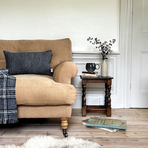 May include: A tan coloured sofa with a dark grey throw pillow and a black and white plaid throw blanket. A small wooden side table with a grey mug, a stack of books, and a vase with dried flowers sits next to the sofa.