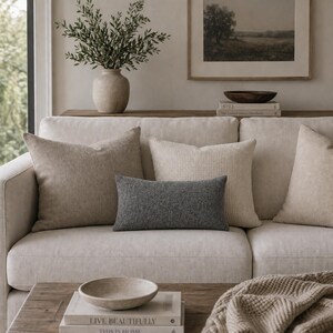 Op de afbeelding: Een neutrale bank met verschillende decoratieve kussens in verschillende texturen en tinten beige en grijs. Een klein, rechthoekig grijs kussen staat in het midden. Een vaas met groen en een ingelijst landschapsschilderij zijn zichtbaar op de achtergrond.