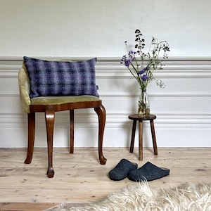 May include: A wooden chair with a green velvet seat and a purple and grey plaid cushion. The chair is in a room with white walls and wooden floors. There is a small wooden stool with a vase of purple flowers on it.