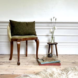 May include: A green velvet cushion sits on a wooden chair with a light green velvet seat. The chair has four legs and is in a room with white walls and wooden floors. A small wooden stool with a stack of books and a ceramic cup sits in the foreground.