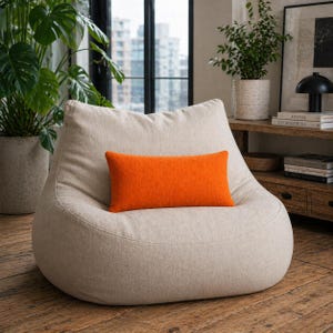 Op de afbeelding: Een beige zitzak met een oranje rechthoekig kussen. De stoel staat in een woonkamer met planten en een houten vloer. Het kussen heeft een levendige oranje kleur en vormt een contrast met de neutrale tinten van de stoel.