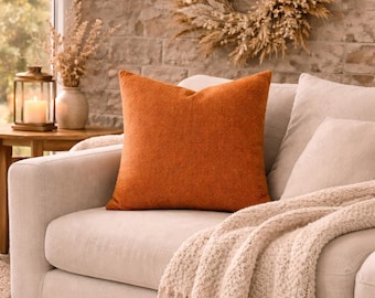May include: An orange textured throw pillow rests on a beige sofa, complemented by a cream-colored throw blanket with fringe. A decorative wreath hangs on the stone wall behind the sofa. A candle lantern and vase with dried flowers are on a wooden side table.
