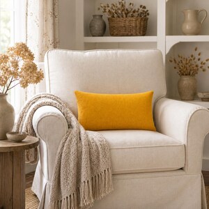 Op de afbeelding: Een comfortabele crèmekleurige fauteuil met een mosterdgeel rechthoekig kussen. Een beige deken met franjes is over de arm gedrapeerd. De kamer is ingericht met droogbloemen in vazen en een kleine houten bijzettafel, wat een warme, uitnodigende sfeer creëert.