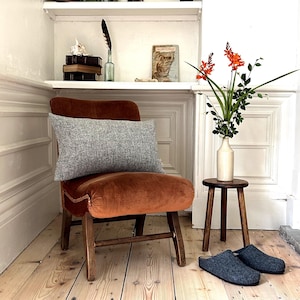 May include: A brown velvet armchair with a grey tweed cushion. The chair is in a room with wooden floors and a small wooden stool with a vase of orange flowers on top.