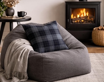 May include: A gray beanbag chair with a plaid pillow and a fringed throw blanket. A black fireplace with a burning fire is in the background. A small black table with a black mug and vase is to the left.