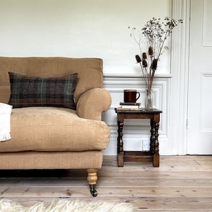 May include: A tan coloured sofa with a plaid throw pillow and a wooden side table with a mug and books on top. The table has a carved design on the legs.