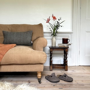 May include: A tan linen sofa with a grey tweed cushion and an orange throw. A small wooden side table with a vase of orange flowers and a stack of books sits next to the sofa. Two grey wool slippers are on the wooden floor.