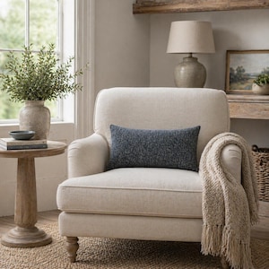 Op de afbeelding: Een crèmekleurige fauteuil met een marineblauw rechthoekig kussen. Een beige deken met franjes is over de arm gedrapeerd. Een kleine houten bijzettafel heeft een vaas met groen en een boek. Een lamp staat op een plank op de achtergrond.