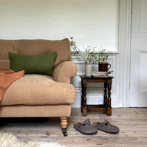 May include: A tan linen sofa with a green throw pillow and an orange throw blanket. A small wooden side table with a vase of white flowers, books, and a mug sits next to the sofa. Two grey wool slippers are on the floor.