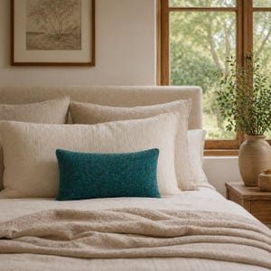 Op de afbeelding: Een bed met verschillende kussens in neutrale tinten, waaronder een rechthoekig turquoise sierkussen. Het bed is opgemaakt met een beige deken en lakens. Een ingelijste foto hangt aan de muur en een vaas met groen staat op een houten nachtkastje.