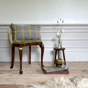 May include: A green velvet chair with a blue and yellow plaid cushion. The chair has four legs and is sitting on a wooden floor. There is a small wooden stool with a brown mug on it, and a stack of books next to the stool.