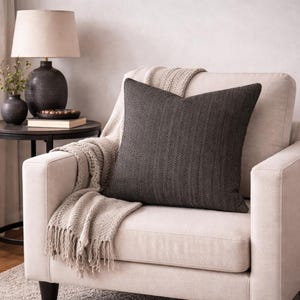 Op de afbeelding: Een donkergrijs decoratief kussen met een visgraatpatroon ligt op een crèmekleurige fauteuil. Een beige sprei met franjes is over de stoel gedrapeerd. Een bijzettafel met een lamp en decoratie staat op de achtergrond.