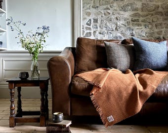 Peut inclure: Un canapé en cuir marron avec une couverture en laine orange brûlée et deux coussins. Un coussin est marron et l'autre est bleu. Le canapé est devant un mur en pierre.