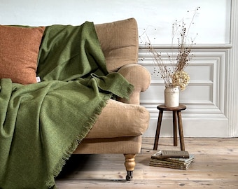 May include: A green throw blanket with fringe edges draped over a tan sofa with a brown pillow. A vase with dried flowers sits on a wooden stool with a stack of books.