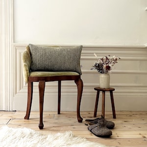 May include: A wooden chair with a green velvet cushion and a grey pillow. The chair is in a room with white walls and wooden floors. There is a small wooden stool with a vase of dried flowers on it.