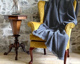 May include: A gray throw blanket draped over a yellow armchair with a floral pattern. The chair has a high back and curved arms. The chair is in front of a stone wall.
