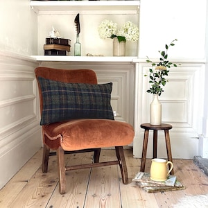 May include: A burnt orange velvet armchair with a plaid lumbar cushion. The chair is in a room with white walls and wooden floors. There is a small wooden stool with a yellow mug on it.