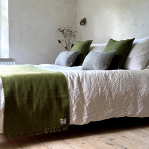 May include: A green throw blanket with fringed edges is draped over a white linen bed with green and grey throw pillows.