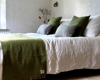 May include: A green throw blanket with fringe edges is draped over a white linen bed with green and gray throw pillows.
