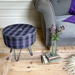 May include: A round, upholstered footstool with a purple and gray plaid pattern. The stool has a gray button in the center and sits on three dark metal hairpin legs. A gray sofa and a small glass bottle with flowers are in the background.