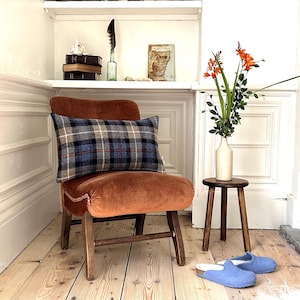 May include: A brown velvet armchair with a plaid lumbar cushion. The chair is in a room with white walls and wooden floors. There is a small wooden stool with a vase of orange flowers on it.