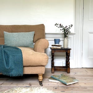 May include: A tan linen sofa with a teal throw blanket and a light blue cushion. A small wooden table with a blue mug and a vase of flowers sits next to the sofa.