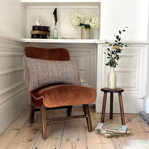 May include: A burnt orange velvet armchair with a tweed lumbar cushion sits on a wooden floor. A small wooden stool with a white ceramic vase holding a sprig of greenery sits next to the chair.