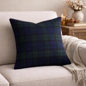 May include: A square cushion featuring a navy blue and green tartan pattern. The cushion is placed on a beige sofa. The design is classic and traditional, suitable for home decor. The cushion is a decorative accent.