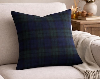 May include: A square pillow with a navy blue and green plaid pattern. The pillow is on a beige sofa. The pillow has a classic, traditional design, suitable for home decor. The pillow is a decorative accent.