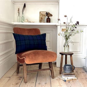 May include: A burnt orange velvet armchair with a dark blue and green plaid lumbar cushion. The chair is in a room with white walls and wooden floors. There is a small wooden stool with a stack of books and a grey mug on top.