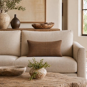 Op de afbeelding: Een beige bank met een bruin en zwart geblokt lendenkussen. Een houten salontafel heeft een kleine, getextureerde vaas met groen. Decoratieve vazen en een houten kom staan op een plank op de achtergrond.