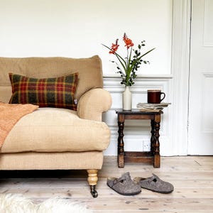 May include: A tan linen sofa with a plaid throw pillow and a burnt orange throw blanket. A small wooden side table with a vase of orange flowers and a mug on top sits next to the sofa. Two grey wool slippers are on the wooden floor.