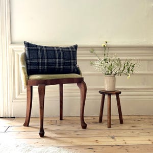 May include: A wooden stool with a green velvet cushion and a plaid pillow. The stool is in front of a white wall and there is a vase of white flowers on a smaller wooden stool.