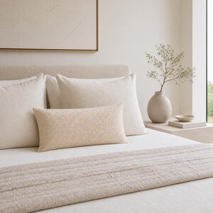 Op de afbeelding: Een bed met een neutraal kleurenpalet, met een beige getextureerd lendenkussen, twee vierkante kussens en een beige bedloper. Een vaas met groen en een schilderij staan op de achtergrond.