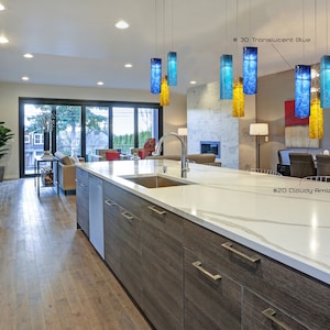 May include: A modern kitchen with a large island featuring a white worktop and wooden cabinets. The island has a stainless steel sink and a bowl of apples on the counter. There are five glass pendant lights hanging above the island, each with a different colour: blue, yellow, and amber. The text "#20 Cloudy Amber", "#21 Blue Lagoon", and "#30 Translucent Blue" is visible in the image.