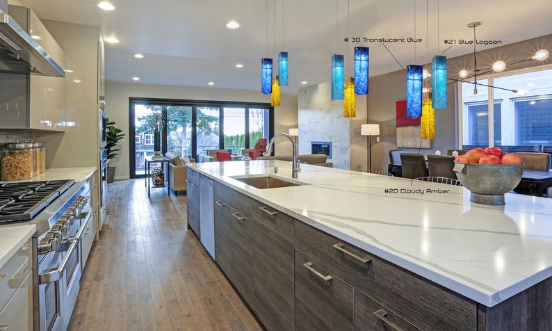 May include: A modern kitchen with a large island featuring a white worktop and wooden cabinets. The island has a stainless steel sink and a bowl of apples on the counter. There are five glass pendant lights hanging above the island, each with a different colour: blue, yellow, and amber. The text "#20 Cloudy Amber", "#21 Blue Lagoon", and "#30 Translucent Blue" is visible in the image.