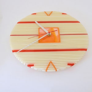May include: A round wall clock with a cream-colored face featuring horizontal lines and red stripes. The clock has an orange square in the center and silver hands. The clock's diameter is approximately 12 inches.