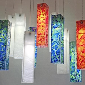 May include: A collection of rectangular pendant lights suspended at varying heights. The fixtures feature a mix of solid white panels and translucent panels with vibrant blue, green, orange, and red abstract patterns, each attached by thin wires.