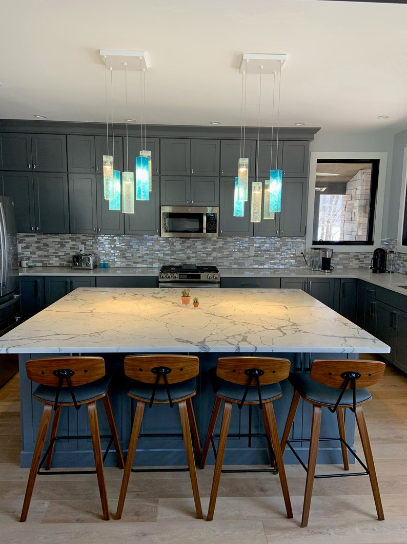 May include: A kitchen island with a white marble worktop and four wooden bar stools. The island is lit by a modern light fixture with blue glass pendants.