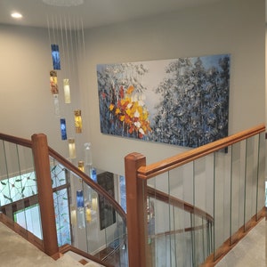 May include: A modern glass chandelier with blue and gold glass panels hangs from the ceiling in a home with a wooden staircase and glass railing. A large abstract painting of a forest scene hangs on the wall behind the staircase.