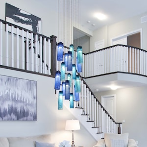 May include: A modern blue glass chandelier with multiple tiers of rectangular glass shades hanging from a white ceiling in a contemporary home with a white staircase.